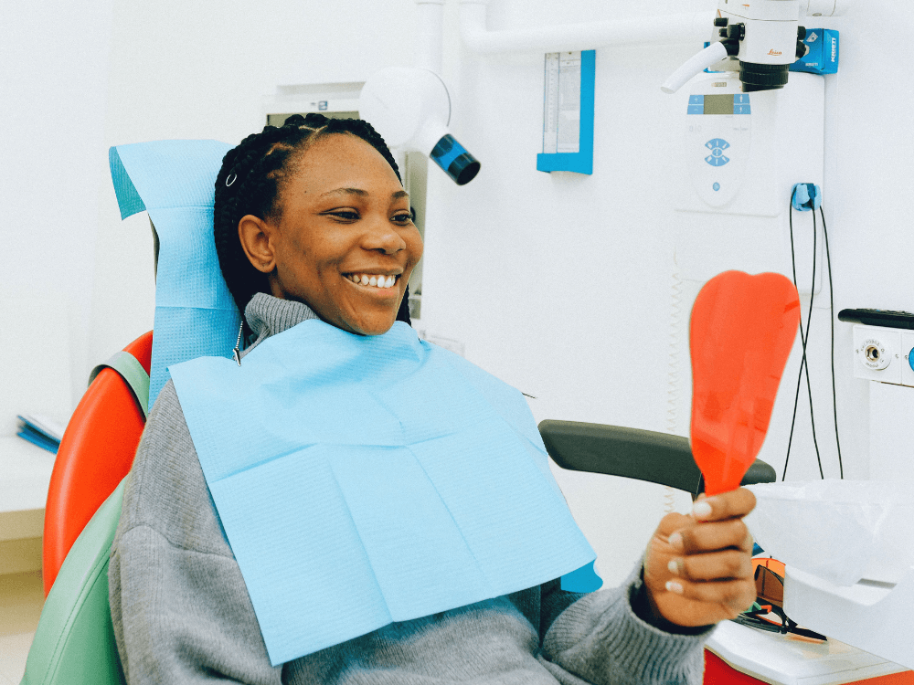 Paciente sentada na cadeira odontológica sorri enquanto segura um espelho de mão para observar os dentes durante a consulta.