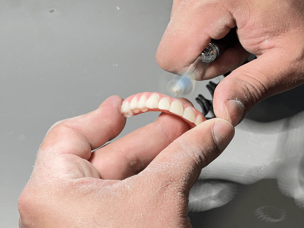 Profissional segura uma prótese dentária e utiliza uma ferramenta rotativa para realizar o acabamento da estrutura protética.