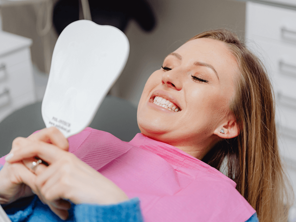 Paciente deitada na cadeira odontológica, sorrindo e observando os dentes em um espelho de mão durante o cuidado com o sorriso. Tudo para explicar prótese e protocolo antes e depois.