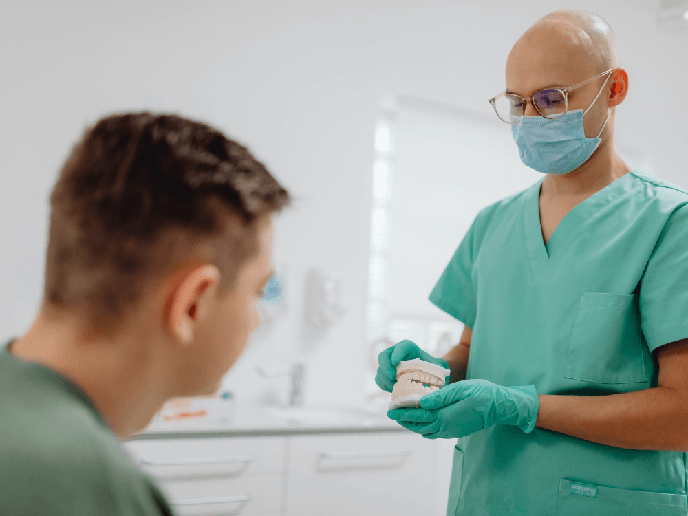 Dentista com máscara, óculos e luvas apresenta um modelo de arcada dentária a um paciente durante uma consulta odontológica.