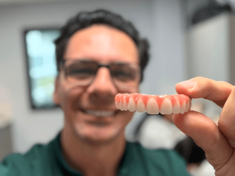 Homem sorridente segura uma prótese dentária com dentes brancos em primeiro plano, ilustrando uma opção de reabilitação oral.