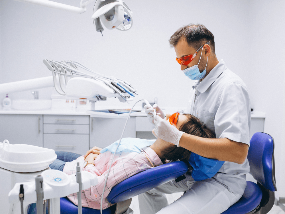 Dentista com máscara, luvas e óculos de proteção atende uma paciente reclinada na cadeira durante uma consulta odontológica. Tudo para explicar os tipos de implante dentário.