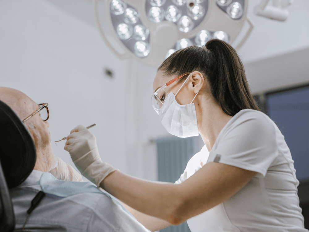Dentista com máscara, luvas e óculos de proteção examina a boca de um paciente idoso durante um atendimento odontológico.